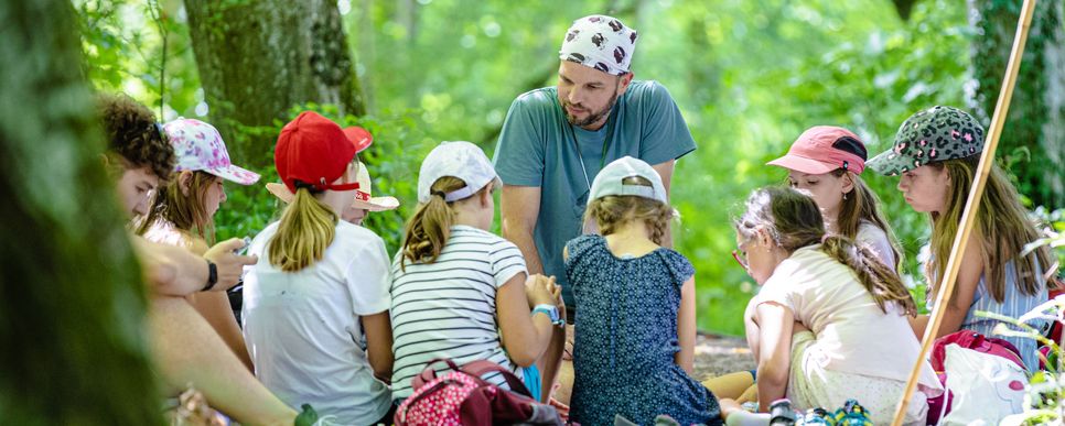 Ein Betreuer erklärt Kindern im Wald etwas.