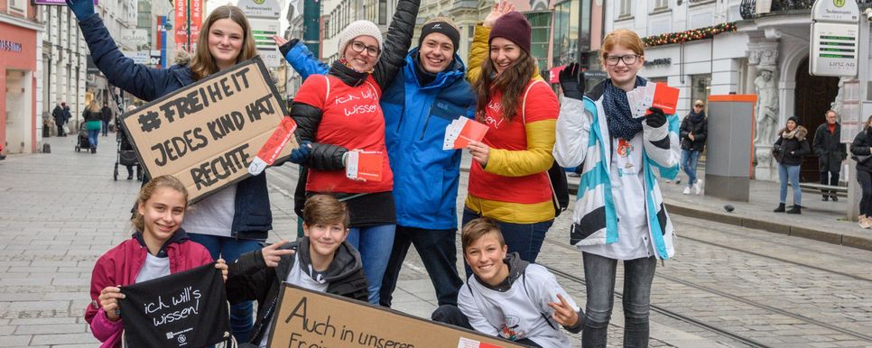 Foto: Haijes Eine fröhliche Gruppe junger Menschen steht in Fußgängerzone. Sie haben ein Schild mit der Aufschrift „JEDES KIND HAT RECHTE“.