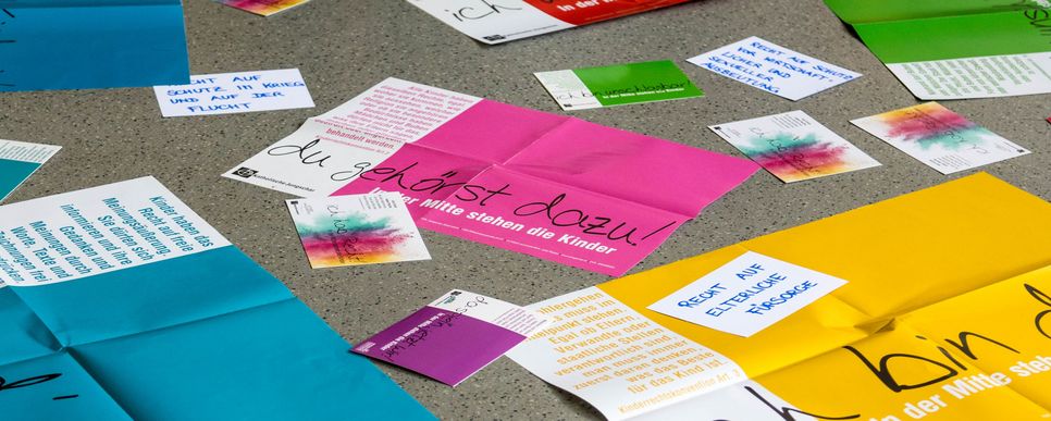 Auf dem Boden liegen bunte Zettel und Flyer, darunter ein pinker mit der Aufschrift "Du gehörst dazu!".