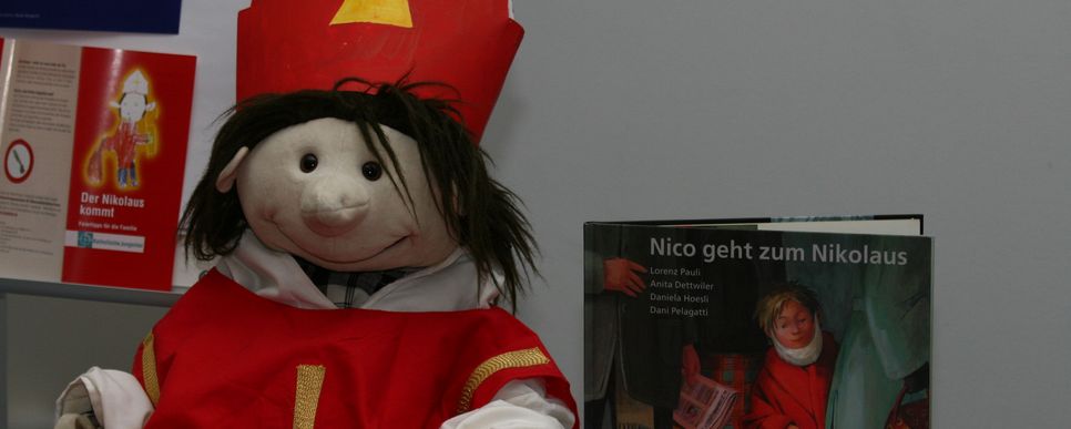 Eine Puppe mit Bischofsmütze und rotem Mantel stellt den heiligen Nikolaus dar. Daneben steht ein Kinderbuch mit dem Titel „Nico geht zum Nikolaus“. Im Hintergrund sind weitere Broschüren zum Thema Nikolaus zu sehen.