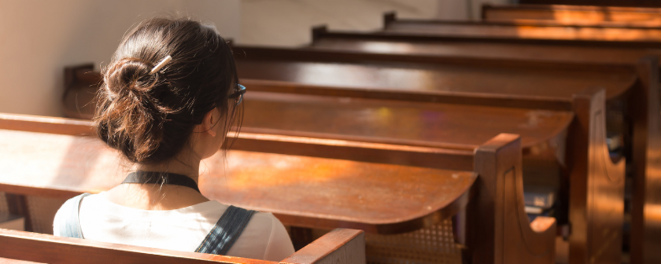 Eine junge Frau sitzt in einer Kirchenbank mit dem Blick nach vorne gerichtet.