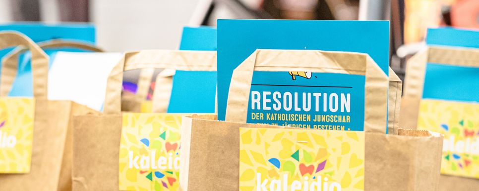 Mehrere Papiertaschen stehen nebeneinander. In jeder Tasche steckt ein türkisfarbener Flyer mit der Aufschrift "Resolution der Katholischen Jungschar".