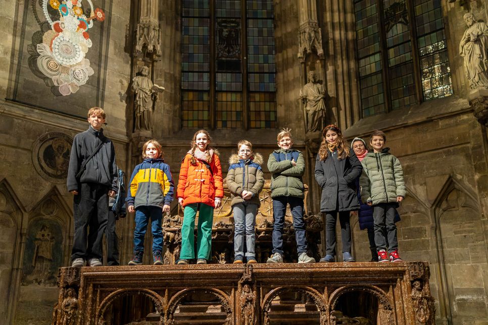 Kinder und Jugendliche stehen auf einem Chorgestühl in einer Kirche. Hinter ihnen sind hohe, bunte Fenster zu sehen.