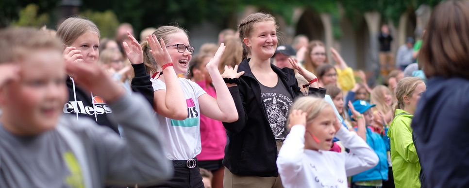 Foto: Hanner/KJSÖ Viele Kinder und Jugendliche stehen im Freien zusammen. Sie lachen und machen mit ihren Händen Bewegungen über dem Kopf, als ob sie an einem Spiel oder einer Aktivität teilnehmen. Die Stimmung wirkt fröhlich und lebhaft.